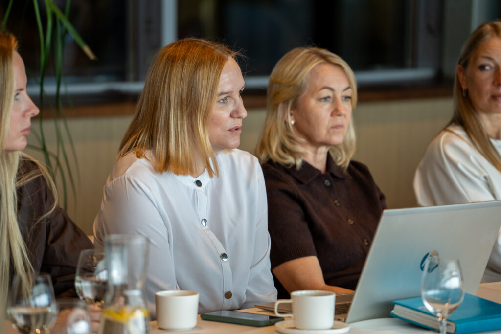 Nuotraukos centre projekto vadovė Svajonė Rainienė sėdi prie stalo. Šalia jos sėdi dar trys moterys. Jos atrodo susitelkusios – Svajonė vilkinti baltus marškinius, kalba arba pristato informaciją, o šalia sėdinti moteris klausosi. Ant stalo yra nešiojamasis kompiuteris, puodeliai, stiklinės su vandeniu ir užrašų knygos – tai rodo, kad vyksta projekto diskusija.
Aplinka atrodo oficiali, tačiau jauki – natūralaus apšvietimo, su augalais fone ir neutraliomis spalvomis. Atmosfera profesionali ir bendradarbiavimo kupina.