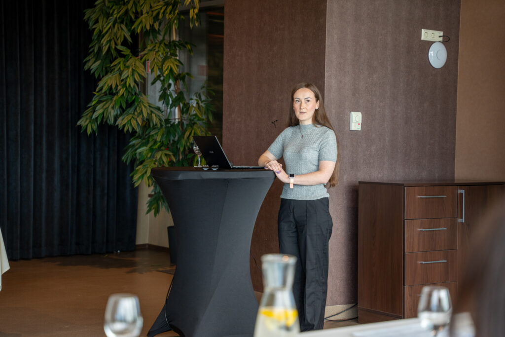 Nuotraukoje matoma pagalbos priiman sprendimus specialistė Vaida Grigelytė-Jonavičienė, stovinti prie aukšto stalo su nešiojamuoju kompiuteriu ir kalbanti ar pristatanti informaciją renginio metu. Ji dėvi pilką megztinį ir juodas kelnes, atrodo susitelkusi ir pasiruošusi pristatymui. Aplinkoje vyrauja neutralios spalvos – tamsiai ruda siena, užuolaidos ir žalios augalų šakos fone. Ant stalo matyti taurė su vandeniu ir kompiuteris, o pirmame plane – kiti susitikimo dalyviai. Atmosfera rimta ir profesionali.