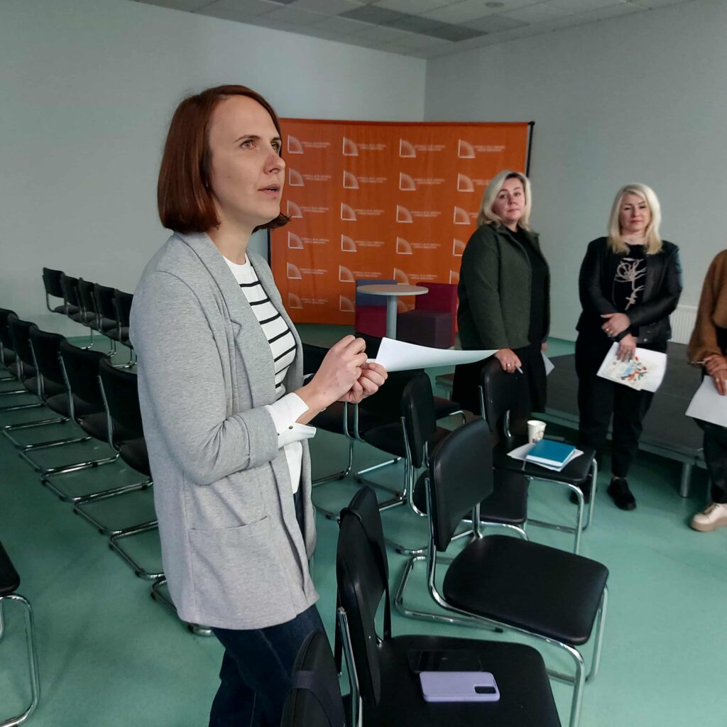 Salėje tarp kėdžių stovi Pagalbos priimant sprendimus paslaugos konsultantė metodininkė Vitalija Kazragienė. Ji kalba ir rankoje laiko baltą popieriaus lapą. Keli dalyviai klausosi jos, fone matyti oranžinis stendas su simbolika.