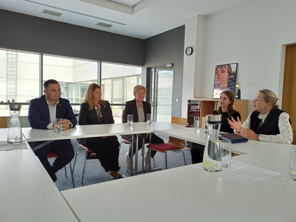 Agentūros salėje, prie didelio stalo sėdi Asmens su negalia teisių apsaugos direktorė Eglė Čaplikienė, Pertvarkos projekto vadovė Svajonė Rainienė, tinklaveikos specialistė Marijona Janavičienė, Šilalės r. meras Tadas Bartkus, vicemerė Jolanta Skrodienė, Agentūros komunikacijos ekspertė Urtė Alksninytė. Direktorė Eglė Čaplikienė kalba, kiti žmonės žiūri į ją ir klauso.