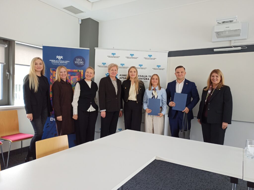 Agentūros salėje, prie didelio stalo stovi Asmens su negalia teisių apsaugos direktorė Eglė Čaplikienė, Pertvarkos projekto vadovė Svajonė Rainienė, tinklaveikos specialistė Marijona Janavičienė, Šilalės r. meras Tadas Bartkus, vicemerė Jolanta Skrodienė, socialinių dirbtuvių „Žadeikių skoniukas“ atstovės Iveta Jokužienė, Vilma Stirbienė, Edita Undaravičienė. 