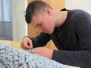 Nuotraukoje matomas jaunas vyras, sėdintis prie stalo ir susikaupęs atliekantis rankdarbius. Jis dėvi tamsiai pilką megztinį su sagutėmis prie kaklo. Ant stalo priešais jį guli šviesiai mėlynas, pūkuotas siūlų audinys, prie kurio jis kruopščiai dirba – galbūt mezga ar pina. Aplinka atrodo švari ir tvarkinga, šviesiame kambaryje su medinėmis durimis ir šviesa, sklindančia pro langą. Bendras įspūdis – ramus, susikaupimo ir kruopštumo momentas.