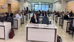 Nuotraukoje pavaizduota grupė žmonių, kurie sėdi prie stalų ir klausosi pranešimo.