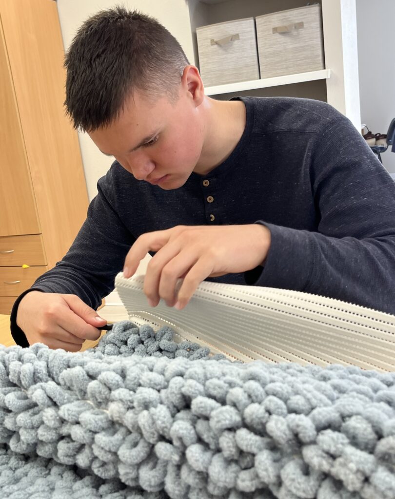 Jaunas vaikinas sėdi prie stalo ir susikaupęs dirba rankomis. Jis dėvi tamsiai pilkus ilgomis rankovėmis marškinėlius su sagutėmis priekyje. Jo dėmesys sutelktas į rankdarbių veiklą – viena ranka laiko baltą plastikinį tinklelį, o kita į jį kruopščiai veria storą, minkštą, šviesiai melsvą siūlą, kuris jau suformavo minkštą, tankų paviršių – panašų į kilimėlį. Fone matyti lentyna su dviem dėžėmis ir medinė spinta. Visa aplinka atrodo tvarkinga ir jauki, o vaikinas – labai susikaupęs ir rimtai įsitraukęs į darbą.