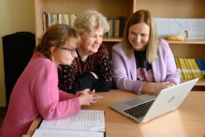 Nuotraukoje yra trys moterys, žiūrinčios į kompiuterį, jos sėdi prie stalo, ant stalo yra keli lapai.