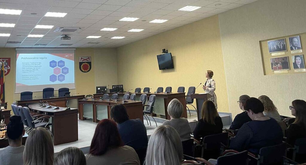 Nuotraukoje matoma posėdžių salė, kurioje vyksta pristatymas. Dešinėje pusėje, prie pranešėjo pulto, stovi Akmenės rajono savivaldybės administracijos Socialinės paramos skyriaus atvejo vadybininkė Simona Ivanauskytė-Jasutė ir kalba auditorijai, žiūrėdama į didelį projekcinį ekraną. Ant ekrano rodomas skaidrės turinys apie psichosocialinę negalią – tekstas ir keli susiję grafiniai elementai.
Auditorijoje sėdi žmonės, daugiausia moterys, stebinčios pranešimą ir atsisukusios į priekį. Priekyje matyti savivaldybės salės įranga: posėdžių stalai su mikrofonais, kėdės, herbai ir vėliavos ant sienų. Aplinka oficiali.