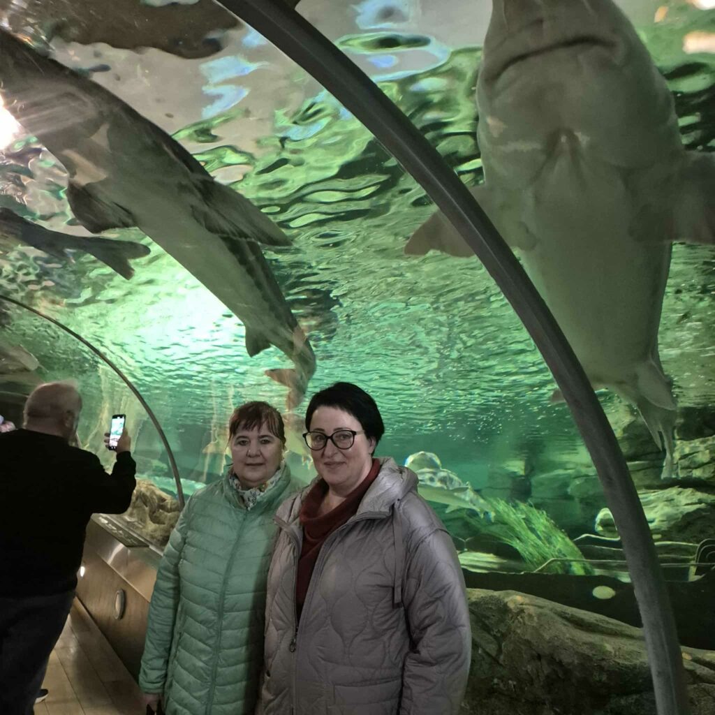 Nuotraukoje matomas stiklinis akvariumo tunelis. Virš galvų, permatomo stiklo kupole, plaukioja du dideli rykliai – jų kūnai masyvūs, šviesiai pilki, ryškiai matosi jų apačia ir judesiai vandenyje. Vanduo virš tunelio žalsvai apšviestas, todėl visas fonas spindi žalsvu atspalviu.
Tunelyje stovi dvi moterys. Jos viena šalia kitos atsisukusios į kamerą. Abi vilki šiltomis, pūstomis striukėmis – viena šviesiai žalsva, kita pilka. Jos šypsosi, atrodydamos ramiai ir jaukiai.
Toliau kairėje pusėje matyti dar vienas žmogus – jis nusisukęs ir savo telefonu fotografuoja ryklius. Grindys medinės, o tunelio pusiau apvalus stiklas tęsiasi abiem kryptimis.