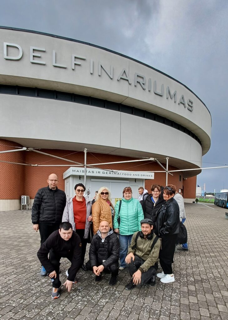 Nuotraukoje matoma žmonių grupė, stovinti lauke prie Klaipėdos delfinariumo pastato. Pagrindiniame fone matosi didelis, apvalus, masyvus pastatas su užrašu „Delfinariumas“ ant viršutinės jo dalies. Pastatas šviesiai pilkas, apačioje – raudonų plytų sienos.
Grupę sudaro devyni žmonės. Šeši jų stovi, o trys – pritūpę priekyje. Visi apsirengę šiltesniais rūbais: striukėmis, paltukais, megztiniais, nes atrodo, kad diena vėsi, dangus apniukęs.
Stovintys žmonės išsirikiavę puslankiu, šypsosi, atrodo gerai nusiteikę. Spalvos įvairios: viena moteris vilki ryškiai žalsvą striukę, kita – pilką, dar kita – juodą odinę striukę. Vyrų striukės daugiausia tamsios.
Pritūpę priekyje trys vyrai. Jie žiūri į kamerą, atrodo ramūs ir susitelkę, vienas iš jų atsirėmęs rankomis į žemę, kiti sėdi ant kulnų.
Aplink – grįstas takas iš pilkų trinkelių, už žmonių matosi nedidelė uždara kiosko tipo patalpa su užrašu „Maistas ir gėrimai“.