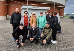 Nuotraukoje matoma žmonių grupė, stovinti lauke prie Klaipėdos delfinariumo pastato. Pagrindiniame fone matosi didelis, apvalus, masyvus pastatas su užrašu „Delfinariumas“ ant viršutinės jo dalies. Pastatas šviesiai pilkas, apačioje – raudonų plytų sienos. Grupę sudaro devyni žmonės. Šeši jų stovi, o trys – pritūpę priekyje. Visi apsirengę šiltesniais rūbais: striukėmis, paltukais, megztiniais, nes atrodo, kad diena vėsi, dangus apniukęs. Stovintys žmonės išsirikiavę puslankiu, šypsosi, atrodo gerai nusiteikę. Spalvos įvairios: viena moteris vilki ryškiai žalsvą striukę, kita – pilką, dar kita – juodą odinę striukę. Vyrų striukės daugiausia tamsios. Pritūpę priekyje trys vyrai. Jie žiūri į kamerą, atrodo ramūs ir susitelkę, vienas iš jų atsirėmęs rankomis į žemę, kiti sėdi ant kulnų. Aplink – grįstas takas iš pilkų trinkelių, už žmonių matosi nedidelė uždara kiosko tipo patalpa su užrašu „Maistas ir gėrimai“.