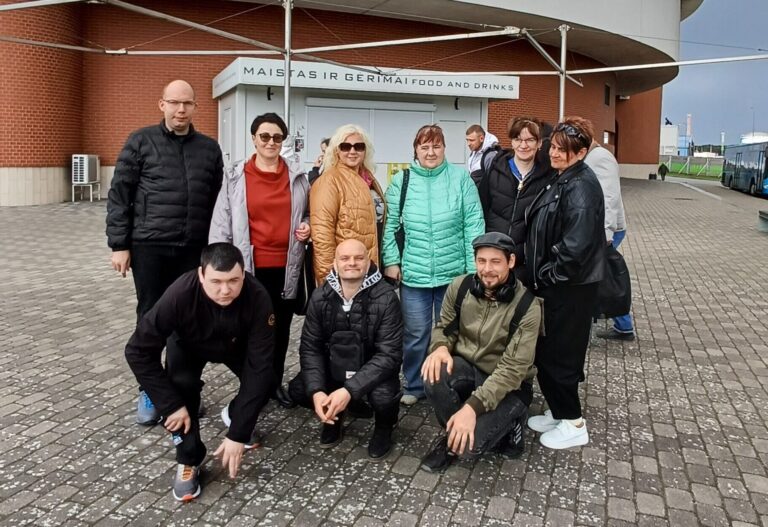 Nuotraukoje matoma žmonių grupė, stovinti lauke prie Klaipėdos delfinariumo pastato. Pagrindiniame fone matosi didelis, apvalus, masyvus pastatas su užrašu „Delfinariumas“ ant viršutinės jo dalies. Pastatas šviesiai pilkas, apačioje – raudonų plytų sienos. Grupę sudaro devyni žmonės. Šeši jų stovi, o trys – pritūpę priekyje. Visi apsirengę šiltesniais rūbais: striukėmis, paltukais, megztiniais, nes atrodo, kad diena vėsi, dangus apniukęs. Stovintys žmonės išsirikiavę puslankiu, šypsosi, atrodo gerai nusiteikę. Spalvos įvairios: viena moteris vilki ryškiai žalsvą striukę, kita – pilką, dar kita – juodą odinę striukę. Vyrų striukės daugiausia tamsios. Pritūpę priekyje trys vyrai. Jie žiūri į kamerą, atrodo ramūs ir susitelkę, vienas iš jų atsirėmęs rankomis į žemę, kiti sėdi ant kulnų. Aplink – grįstas takas iš pilkų trinkelių, už žmonių matosi nedidelė uždara kiosko tipo patalpa su užrašu „Maistas ir gėrimai“.