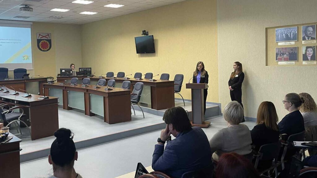 Nuotraukoje matoma savivaldybės posėdžių salė, kurioje vyksta pristatymas. Dešinėje pusėje prie pranešėjo pulto stovi Akmenės rajono socialinių paslaugų namų socialinių dirbtuvių socialinė darbuotoja Enrika Klusienė ir rinkodaros vadybininkė Lidija Jatkauskė ir kalba auditorijai. Priekyje, auditorijoje, sėdi klausytojai, o vienas vyras pirmoje eilėje atsirėmęs ranka į smakrą klausosi ypač įdėmiai. Kairėje pusėje ant projekcinio ekrano matoma skaidrė su tekstu ir logotipu, tačiau tik fragmentiškai. Ant sienos kabo keli herbai ir nuotraukų rėmai, pabrėžiantys oficialią institucijos aplinką.