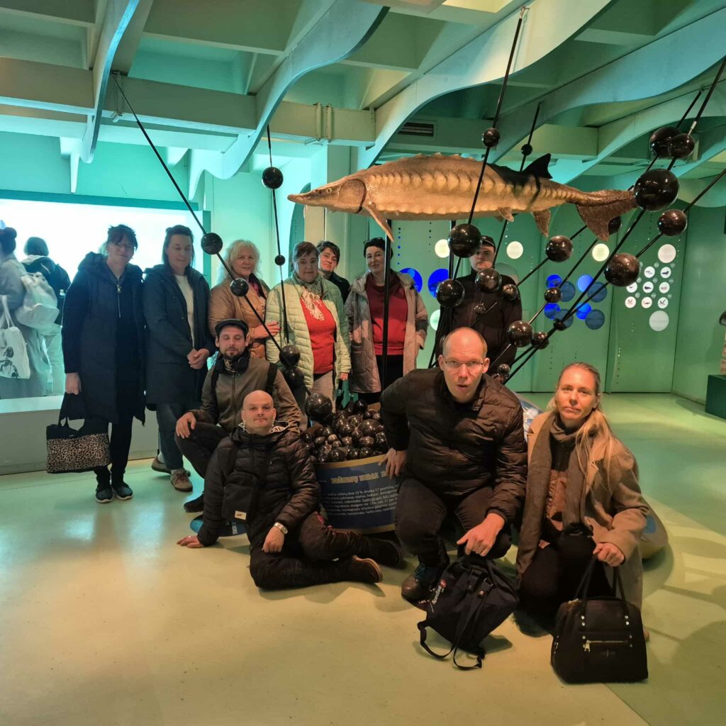 Nuotraukoje matoma grupė žmonių, nusifotografavusių jūrų muziejaus ekspozicijoje. Visi jie stovi ar tupi erdvioje, žalsvai apšviestoje patalpoje, primenančioje modernų akvariumą ar mokslinę galeriją.
Grupėje yra apie vienuolika žmonių. Dalis jų stovi gale. Trys vyrai ir viena moteris – pritūpę ar atsisėdę priekyje. Visi apsirengę šiltais paltais ar striukėmis. Išraiškos ramios, draugiškos, žmonės atrodo susitelkę ir pasirengę bendram kadrui.
Patalpos centre, už grupės, yra didelė ekspozicinė konstrukcija – ant juodų metalinių strypų pakabintas didelis žuvies muliažas. Tai ilga, siaura žuvis, primenanti eršketą: jos kūnas tamsus, viršus pilkšvas, paviršius šiurkštus, su iškiliais šarvais. Aplink ją pritvirtinta daug juodų, apvalių rutulių, kurie atrodo kaip dekoratyviniai ekspozicijos elementai.
Fone matosi žalsvai melsvos sienos su apvaliomis šviesomis ir langeliais, o kairėje — didelis stiklas, pro kurį matyti kiti lankytojai.