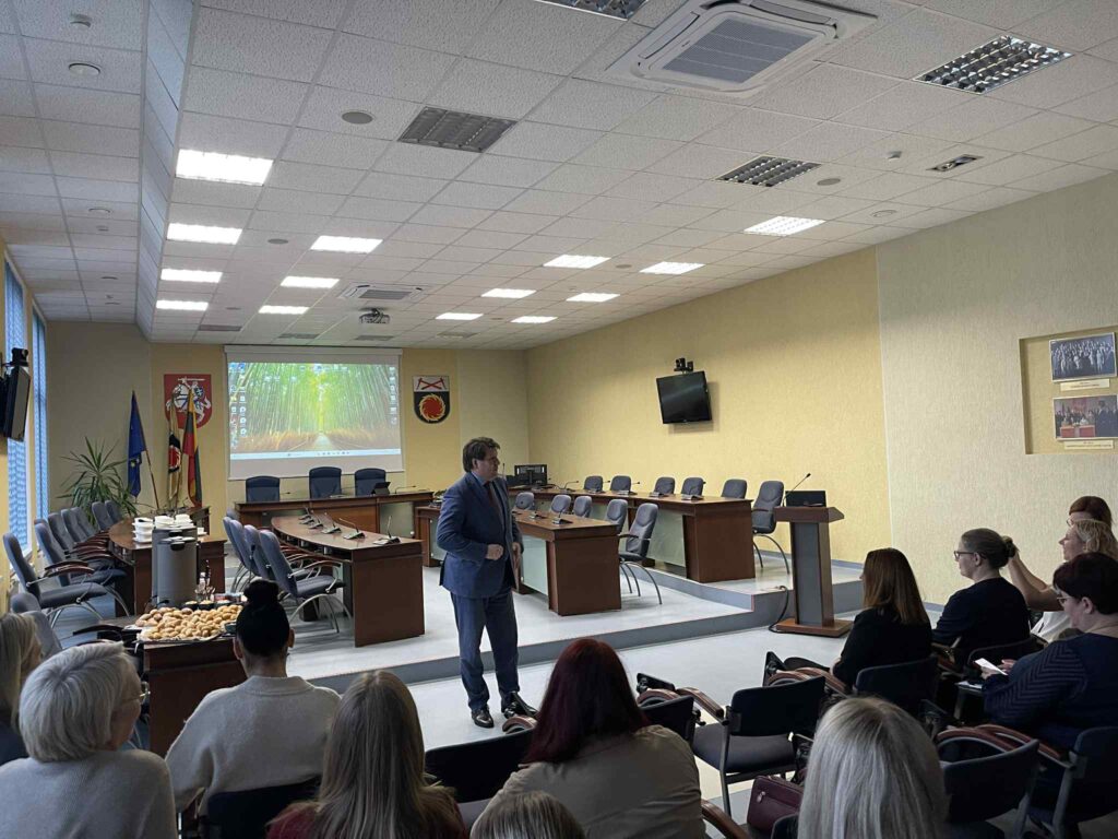 Nuotraukoje Akmenės rajono savivaldybės meras Vitalijus Mitrofanovas. Jis stovi posėdžių salės priekyje prieš susirinkusius dalyvius, vilkėdamas mėlyną kostiumą. Auditorijoje sėdi žmonės, daugiausia moterys, stebinčios jo kalbą ar pristatymą.