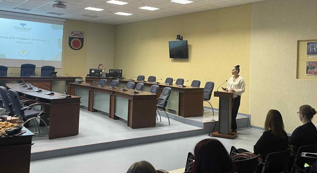 Nuotraukoje matoma savivaldybės posėdžių salė, kurioje vyksta pristatymas. Dešinėje pusėje, prie pranešėjo pulto, stoviAgentūros specialistė Šiaulių miestui ir rajonui Viktorija Krivickaitė-Kriščiotaitė. Moteris vilki šviesų megztinį ir juodas kelnes. Ji kalba auditorijai ir rankoje laiko pultelį skaidrėms valdyti.