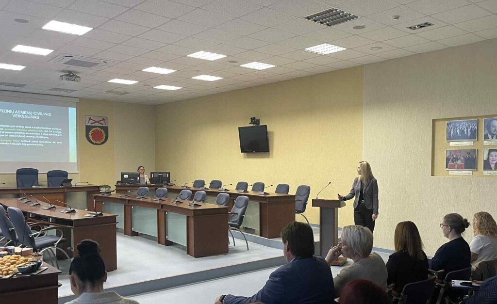 Nuotraukoje matoma savivaldybės posėdžių salė, kurioje vyksta pristatymas. Dešinėje prie pranešėjo pulto stovi Agentūros veiksnumo apribojimo prevencijos specialistė Sandra Grosienė, kalbanti auditorijai. Kairėje pusėje rodomas projekcinis ekranas su skaidre apie fizinių asmenų civilinį veiksnumą. Prie kompiuterių sėdi dar viena moteris, valdanti techniką.