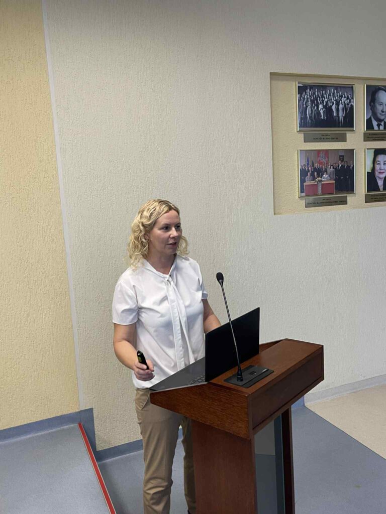 Šioje nuotraukoje Asmens su negalia teisių apsaugos agentūros tinklaveikos specialistė Eglė Jurkutė, stovinti prie medinio pranešėjo pulto. Ji vilki baltus trumpomis rankovėmis marškinius ir smėlio spalvos kelnes. Fone – šviesi sienelė su įrėmintomis nuotraukomis ir portretais, greičiausiai susijusiais su institucijos istorija ar vadovais. Aplinka atrodo oficiali, panaši į savivaldybės ar įstaigos salę.