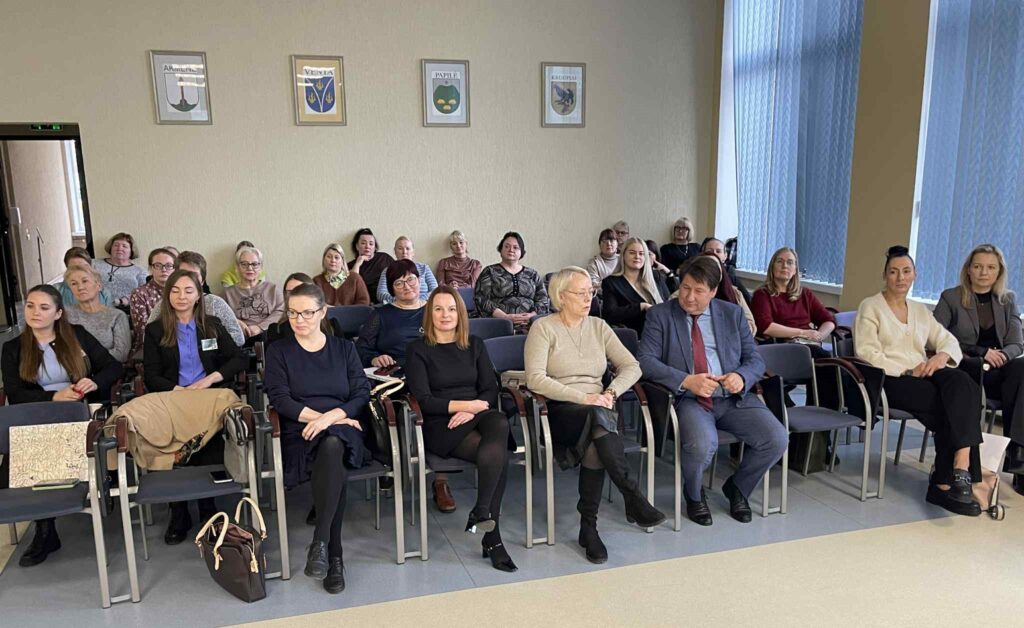 Nuotraukoje matoma vidutinio dydžio salė, kurioje susėdę žmonės. Žmonės sėdi eilėmis išdėstytose kėdėse, nukreipę žvilgsnį į fotografą. Patalpa šviesi, su dideliais langais dešinėje pusėje, pro kuriuos sklinda dienos šviesa. Ant sienos virš galinių eilių kabo keli herbiniai simboliai ar miesto seniūnijų/įstaigos ženklai. Dauguma – suaugusios moterys, skirtingo amžiaus, nuo jaunų iki vyresnių.