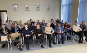 Nuotraukoje matoma vidutinio dydžio salė, kurioje susėdę žmonės. Žmonės sėdi eilėmis išdėstytose kėdėse, nukreipę žvilgsnį į fotografą. Patalpa šviesi, su dideliais langais dešinėje pusėje, pro kuriuos sklinda dienos šviesa. Ant sienos virš galinių eilių kabo keli herbiniai simboliai ar miesto seniūnijų/įstaigos ženklai. Dauguma – suaugusios moterys, skirtingo amžiaus, nuo jaunų iki vyresnių.