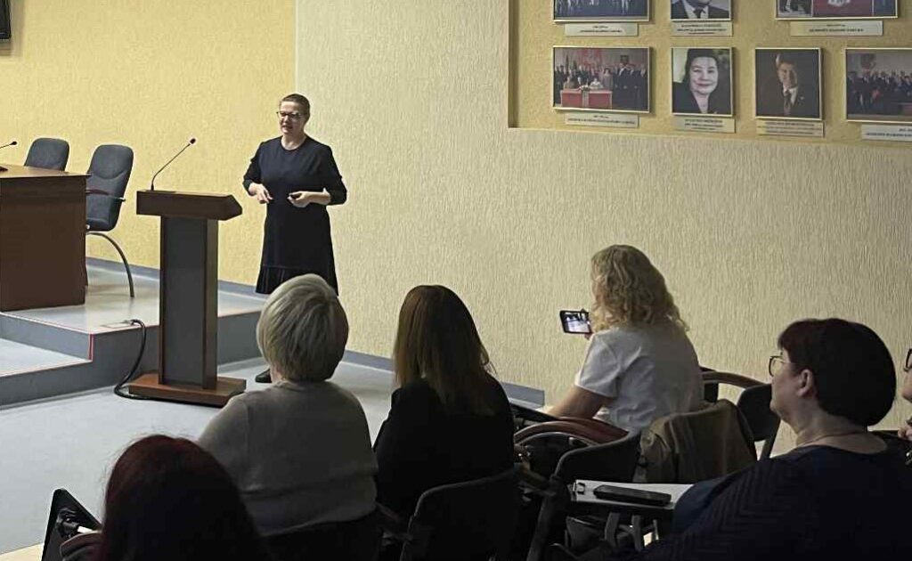 Nuotraukoje matoma Agentūros konsultantė-metodininkė (apsaugotam būstui ir grupinio gyvenimo namams) Donata Šimkutė-Špėgienė, stovinti savivaldybės salėje prie pranešėjo pulto. Ji vilki tamsų, paprastą drabužį ir kalbėdama gestikuliuoja, tarsi aiškintų ar akcentuotų svarbią mintį. Priekyje prieš ją sėdi auditorija – daugiausia moterys, įdėmiai klausosi. Viena moteris dešinėje pusėje laiko telefoną ir, panašu, filmuoja pranešimą.