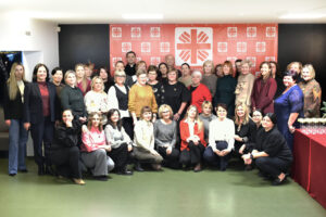 Didelė žmonių grupė – daugiausia moterys – sustoję ir susėdę bendrai nuotraukai patalpoje. Fone matyti raudonas „Caritas“ stendas su logotipais. Visi žiūri į kamerą ir šypsosi, atmosfera šventiška. Dešinėje pusėje matyti stalas su taurėmis.