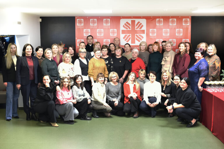 Didelė žmonių grupė – daugiausia moterys – sustoję ir susėdę bendrai nuotraukai patalpoje. Fone matyti raudonas „Caritas“ stendas su logotipais. Visi žiūri į kamerą ir šypsosi, atmosfera šventiška. Dešinėje pusėje matyti stalas su taurėmis.