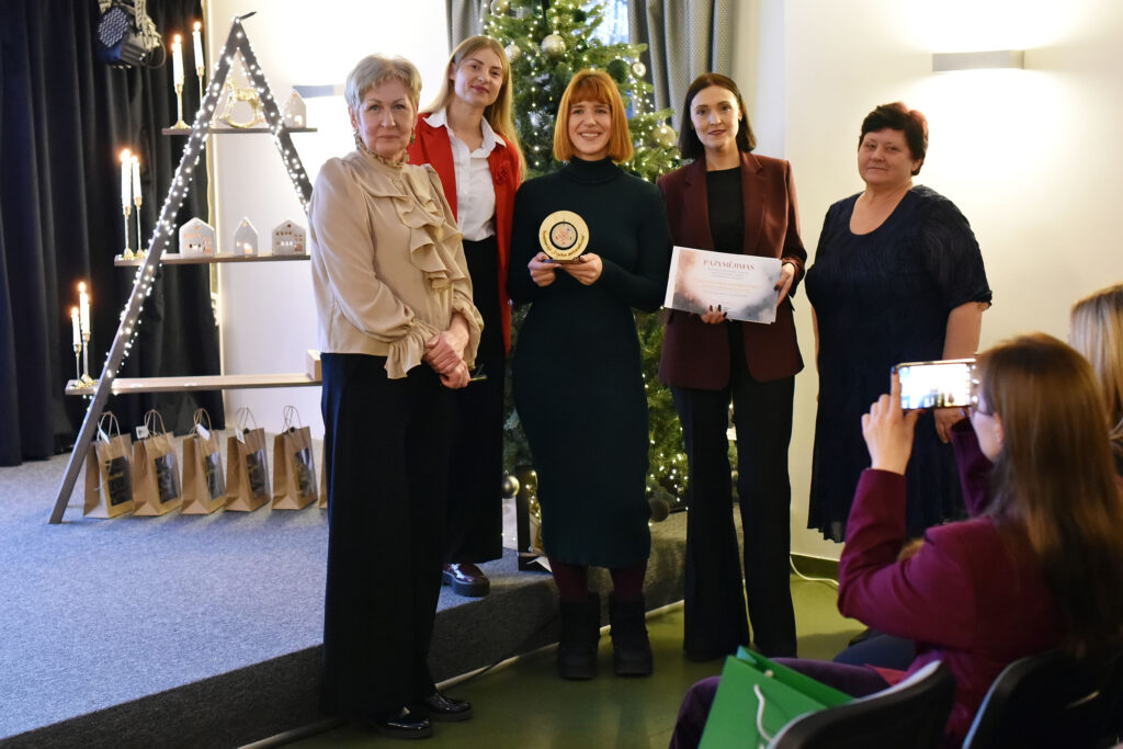 Nuotraukoje matyti grupė moterų, sustojusių pozuoti prie kalėdinės eglės apdovanojimų įteikimo metu. Centre esančios moterys rankose laiko apdovanojimą ir pažymėjimą. Jos šypsosi, atrodo pakiliai. Šalia jų stovi dar kelios renginio dalyvės, taip pat pozuojančios kamerai. Kairėje matyti dekoratyvi trikampė lentyna su žvakėmis ir mažais šviečiančiais nameliais, o apačioje – dovanų maišeliai. Priekyje – žiūrovė, fotografuojanti momentą telefonu.