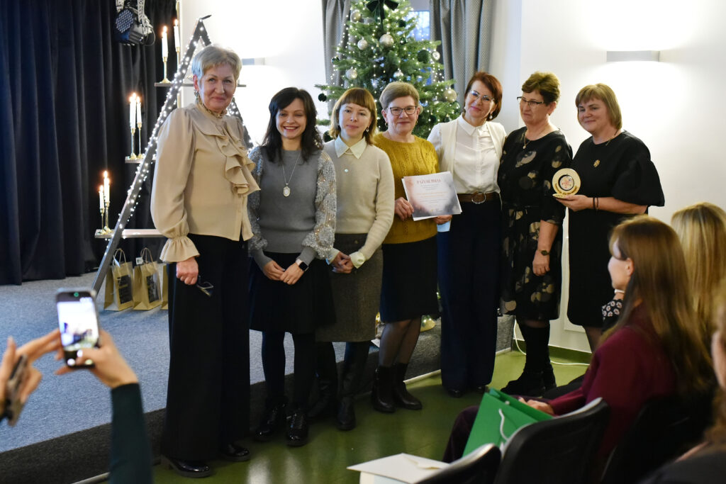 Nuotraukoje grupė moterų pozuoja apdovanojimų įteikimo akimirkai prie kalėdinės eglės. Kelios moterys priekyje laiko padėką ir medinį apdovanojimą. Kitos stovi šalia, šypsosi ir žiūri į kamerą. Atmosfera šilta ir šventiška. Kairėje matoma dekoratyvi trikampė lentyna su uždegtomis žvakėmis ir dovanų maišeliais apačioje. Priekyje kadro – žiūrovės ranka, fotografuojanti šią akimirką telefonu.