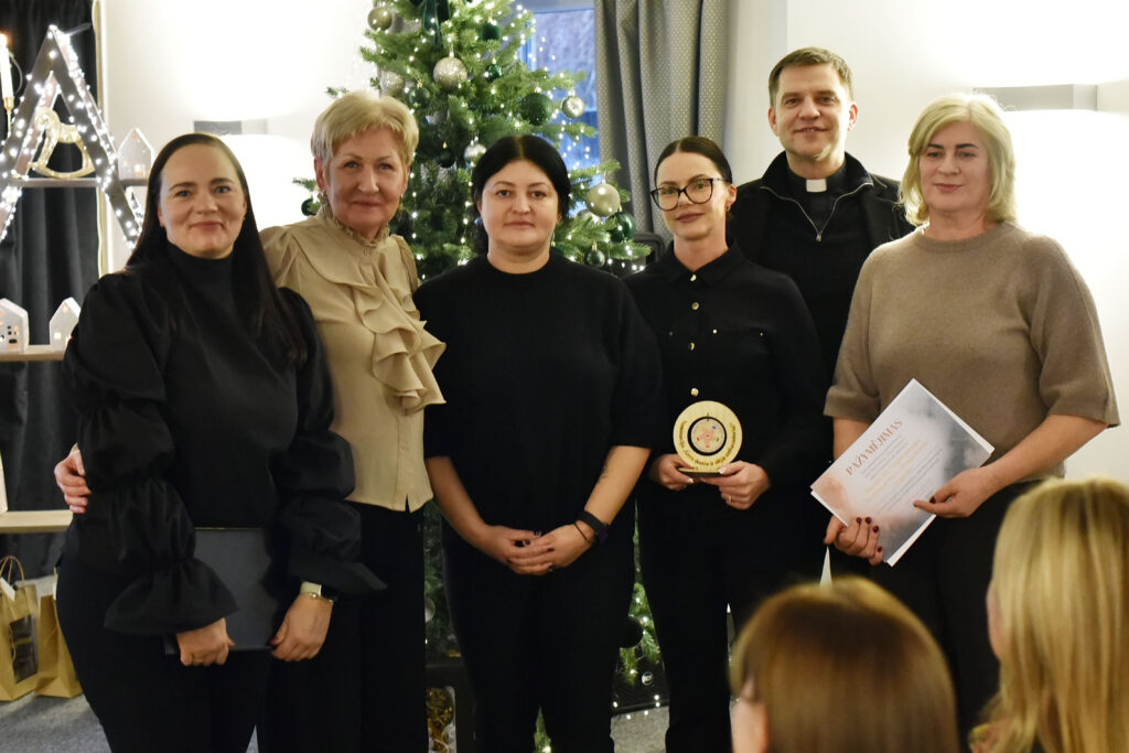 Nuotraukoje matyti grupė žmonių, pozuojančių prie kalėdinės eglės apdovanojimų įteikimo metu. Priekyje stovi kelios moterys – jos šypsosi, laiko apdovanojimą ir pažymėjimą. Greta jų stovi vyras juodais dvasininko drabužiais. Visi dalyviai sustojo draugiškai, artimai vieni prie kitų. Kairėje matoma šventinė dekoracija: trikampė lentyna su žvakėmis ir mažais šviečiančiais nameliais. Fone – eglutė su lemputėmis. Priekyje iš apačios matosi žiūrovų galvos, stebinčios įteikimo momentą.