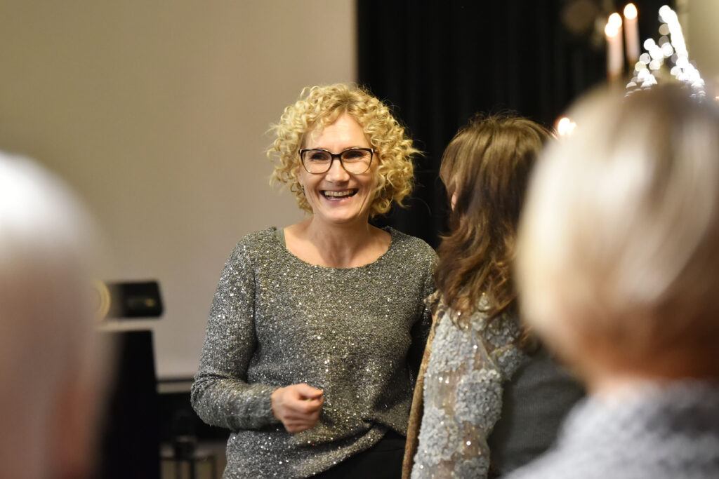 Nuotraukoje matyti šviesiaplaukė moteris su garbanotais plaukais ir akiniais. Ji šypsosi ir bendrauja su kita moterimi, kuri matosi tik iš nugaros. Abi atrodo įsitraukusios į pokalbį, atmosfera lengva ir šventiška. Priekyje matosi kelių žiūrovų siluetai.