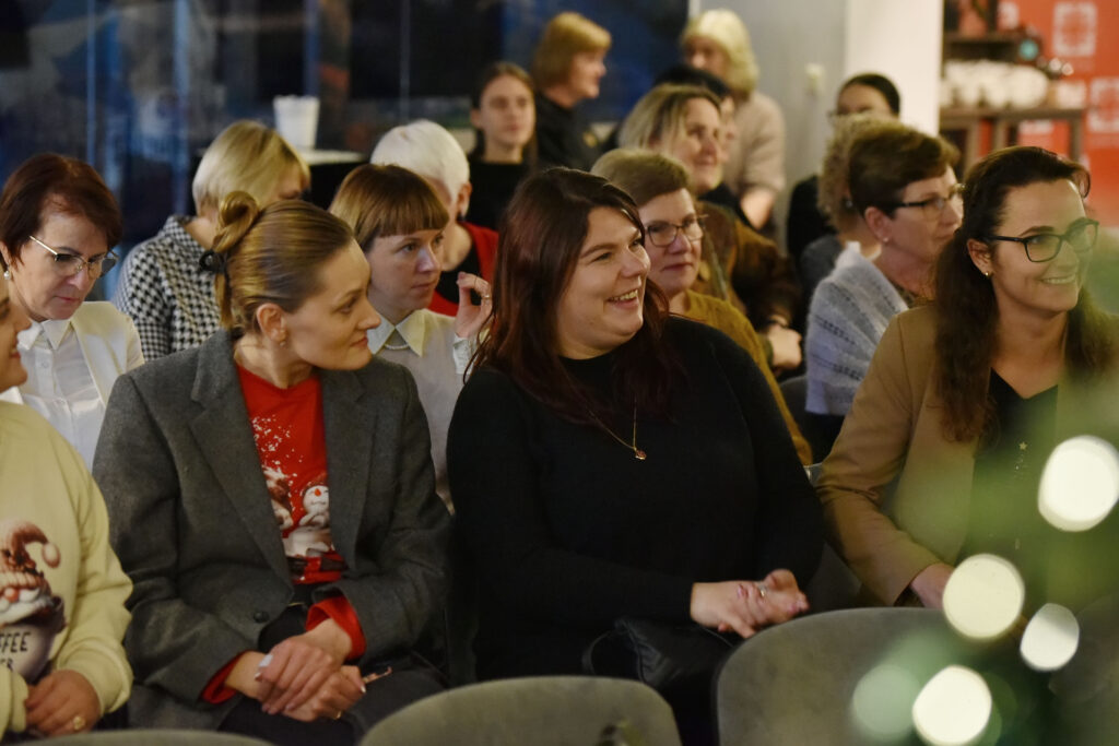 Nuotraukoje matyti auditorijoje sėdinčios renginio dalyvės. Priekiniuose suoluose kelios moterys šypsosi, bendrauja tarpusavyje, atrodo įsitraukusios ir geros nuotaikos. Viena vilki pilką švarką ir raudoną megztinį, kita – juodą megztinį, dar kita – smėlio spalvos švarką. Už jų matyti daugiau sėdinčių dalyvių, kai kurios klausosi, kitos šnekučiuojasi tarpusavyje. Aplinka šilta ir draugiška.