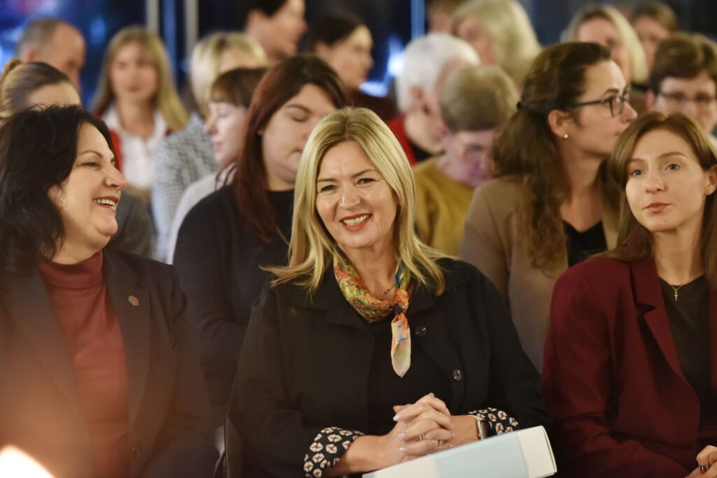 Nuotraukoje matyti auditorijoje sėdinčios renginio dalyvės. Priekyje trys moterys sėdi greta:
Kairėje moteris juodu švarku juokiasi, žiūrėdama į šalia sėdinčią.
Centre moteris šviesiais plaukais, vilkinti juodą švarką ir spalvingą šaliką, šypsosi tiesiai į kamerą.
Dešinėje jauna moteris bordo švarku žiūri į šoną.
Fone matyti daugiau sėdinčių dalyvių, kurios klausosi ar šnekučiuojasi. Atmosfera šilta ir gyva.