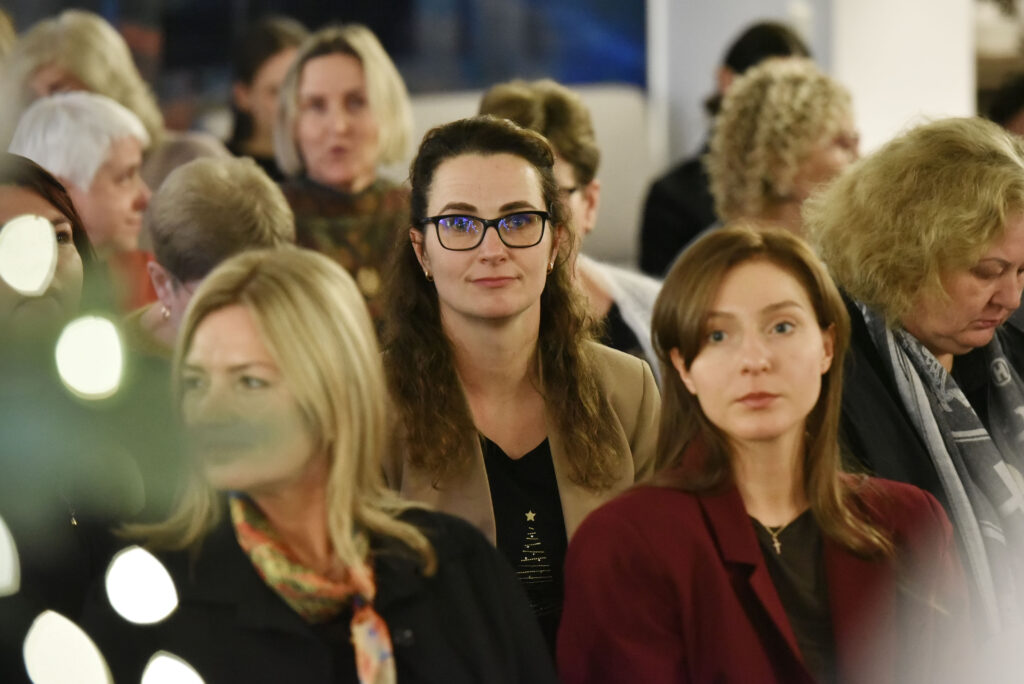 Nuotraukoje matyti renginio dalyvės, sėdinčios auditorijoje. Centre – moteris su akiniais ir šviesiai rudu švarku, žiūrinti tiesiai į kamerą. Šalia jos – jauna moteris bordo spalvos švarku, žiūrinti kiek į šoną. Priekyje kairėje matosi dar viena moteris, tačiau jos veidą dalinai užstoja šventinės lemputės. Fone – daugiau sėdinčių dalyvių, besiklausančių ar bendraujančių tarpusavyje. Atmosfera rami, susitelkusi.