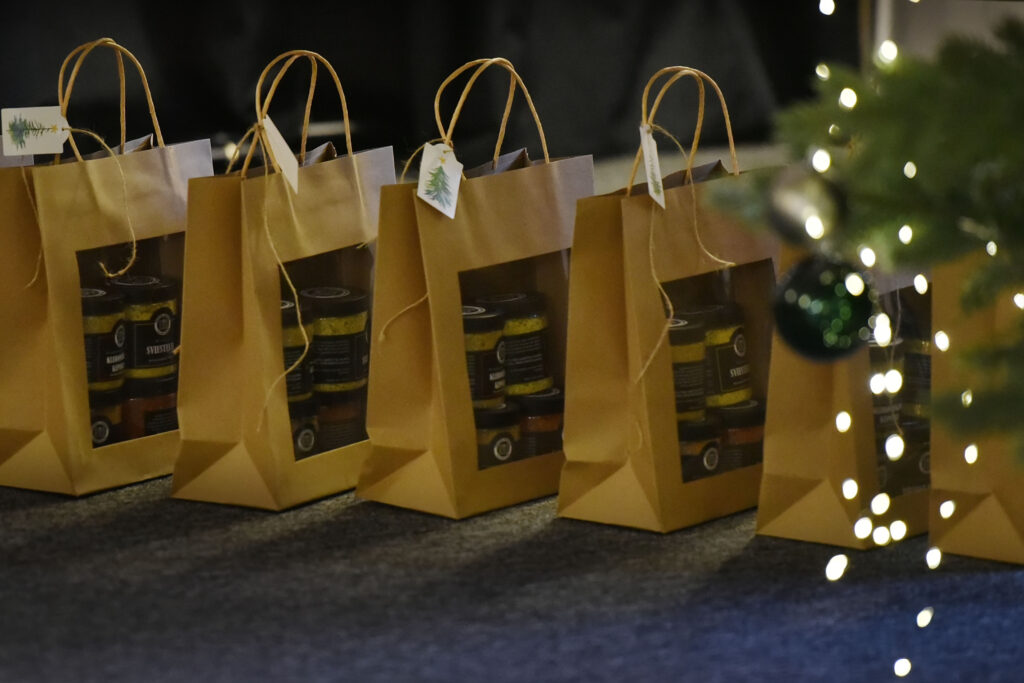 Nuotraukoje matyti eilė popierinių dovanų maišelių, pastatytų ant grindų šalia kalėdinės eglės. Maišeliai rudi, su langeliu priekyje, per kurį matosi viduje sudėti stiklainėliai su maisto gaminiais. Ant kiekvieno maišelio pritvirtinta maža kortelė su nupiešta eglute. Dešinėje kadro pusėje matyti šventinės eglutės šakos ir mirgančios lemputės.