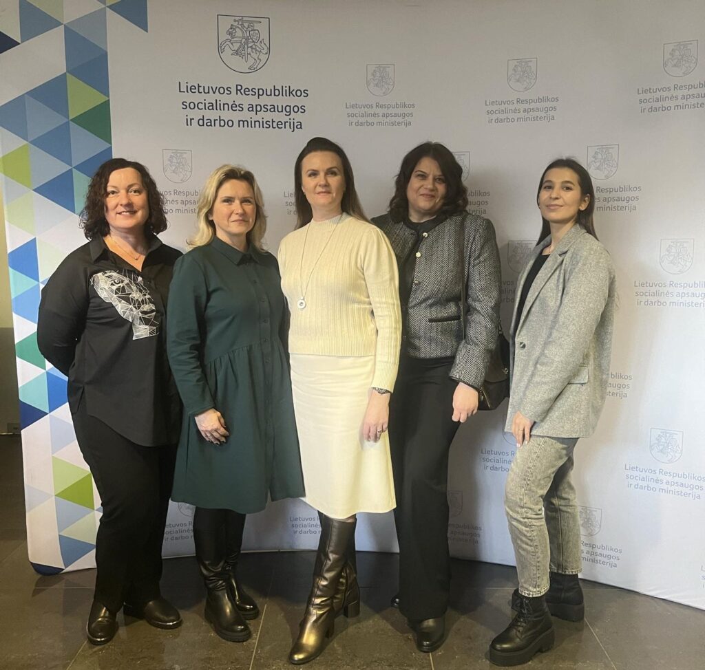 Nuotraukoje matyti penkios moterys, stovinčios greta ir pozuojančios patalpoje. Jos vilki tvarkingus, dalykinius drabužius, atrodo ramios ir savimi pasitikinčios. Už jų – pastatoma siena su oficialiais Lietuvos Respublikos socialinės apsaugos ir darbo ministerijos logotipais.
