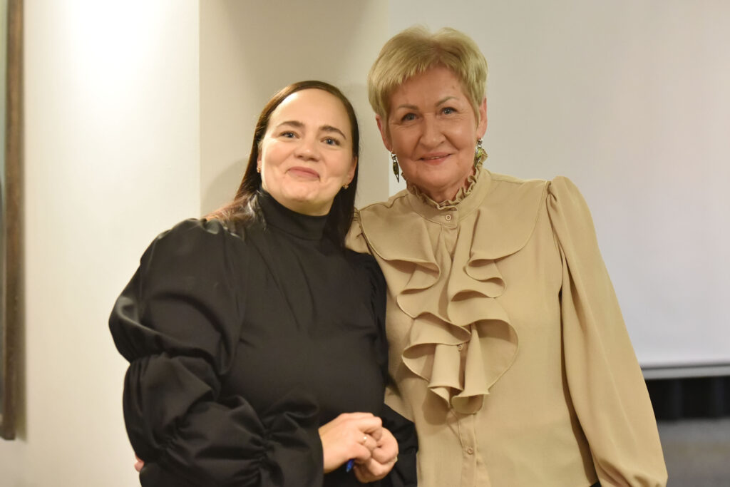 Nuotraukoje dvi moterys stovi greta ir šypsosi kamerai. Kairėje – tamsiaplaukė moteris juodu, pūstomis rankovėmis puoštu drabužiu. Dešinėje – šviesiaplaukė moteris šviesios smėlio spalvos palaidine su dekoratyviais raukiniais. Abi atrodo draugiškai, pozuoja šiltai apsikabinusios per pečius.