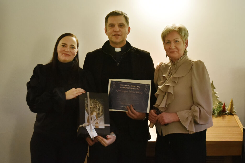 Nuotraukoje stovi trys žmonės:
Kairėje – tamsiaplaukė moteris, vilkinti juodą drabužį su pūstomis rankovėmis. Ji šypsosi ir rankose laiko dovanų dėžutę perrištą kaspinu.
Viduryje – vyras su juoda apranga ir kolaro apykakle (dvasininkas). Jis rankose laiko padėkos ar nominacijos diplomą.
Dešinėje – šviesiaplaukė moteris šviesia palaidine, kuri puošta ryškiais raukiniais priekyje. Ji ramiai pozuoja šalia.
Fone matosi kalėdinės dekoracijos ant stalo.