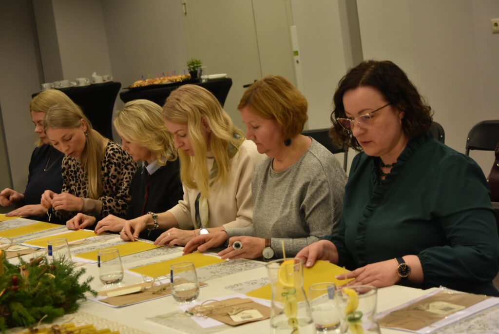 Nuotraukoje matyti šešios moterys, sėdinčios prie ilgo stalo patalpoje. Jos susikaupusios rankomis liečia ir tyrinėja ant stalo padėtus geltonus vaško lakštus. Ant stalo yra stiklinės su vandeniu, popieriai, smulkūs daiktai. Aplinka rami, darbinė.