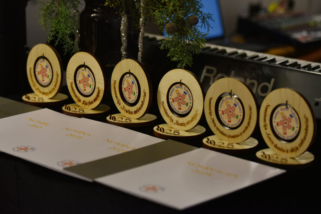 Nuotraukoje ant stalo išdėlioti apdovanojimai ir diplomai. Priekyje matomos baltos aplankų formos padėkos su auksiniu užrašu „Nominacija 2025“. Už jų stovi mediniai apdovanojimai – apvalūs stoveliai su spalvotu socialinių dirbtuvių logotipu centre. Ant jų užrašytos nominacijų kategorijos, pvz. „Kūrybos mazgas“, „Tvarkos ambasadoriai“, „Skonių atradėjai“ ir kt. Fone matosi muzikos instrumentas – klavišinis sintezatorius.