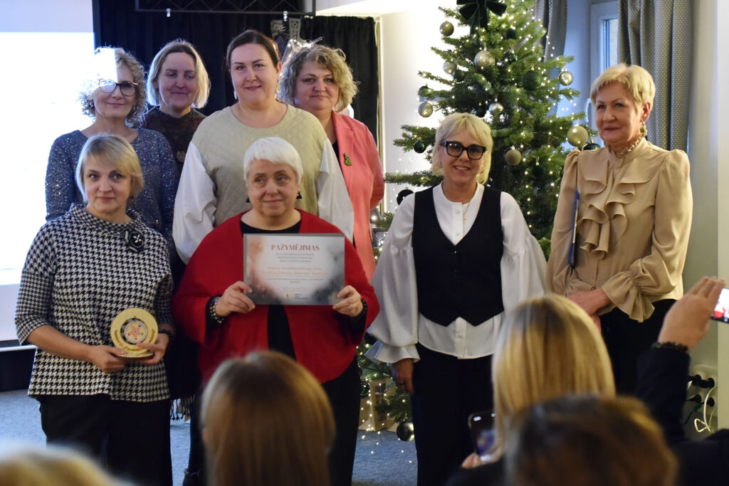 Nuotraukoje matyti grupė moterų, sustojusių prie kalėdinės eglės per apdovanojimų įteikimą. Priekyje kelios moterys laiko padėkas ir medinius apdovanojimus, šypsosi į kamerą. Už jų stovi dar kelios renginio dalyvės, taip pat pozuojančios šiam momentui. Dešinėje pusėje stovi moteris šviesia palaidine su raukiniais.

