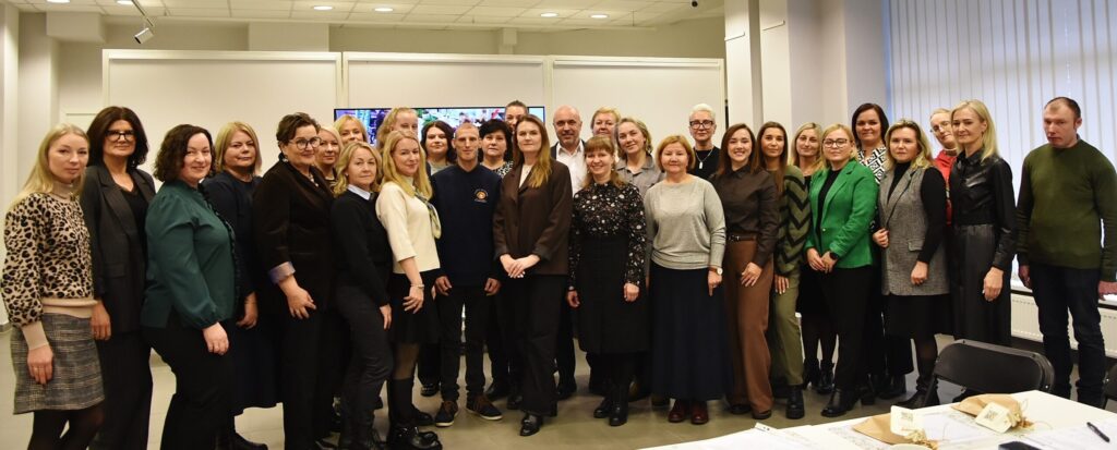 Nuotraukoje – didelė žmonių grupė (apie 30 asmenų), sustojusių kartu patalpoje. Jie stovi keliomis eilėmis, žiūri į kamerą ir pozuoja bendrai nuotraukai. Dauguma apsirengę tvarkingais, dalykiniais ar kasdieniais drabužiais. Aplinka šviesi, už nugaros matyti sienos ir ekranas, atmosfera rami ir oficiali.