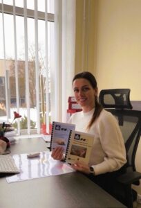 Nuotraukoje matoma moteris, sėdinti prie darbo stalo biuro aplinkoje. Ji sėdi patogioje biuro kėdėje, pasisukusi šiek tiek į fotoaparato pusę, ir švelniai šypsosi. Moteris vilki šviesų, ilgomis rankovėmis megztinį, jos tamsūs plaukai surišti atgal. Rankose ji laiko dvi nedideles lengvai suprantamos kalbos metodines knygeles su spalvotais viršeliais – viena mėlyna, kita gelsva. Ant viršelių matyti iliustracijos ir tekstas. Ant stalo prieš ją yra kompiuterio klaviatūra, pelė, dokumentai ir keli smulkūs biuro daiktai. Už nugaros – didelis langas su vertikaliomis žaliuzėmis; pro langą matosi žiemos vaizdas lauke: sniegas ant žemės, medžiai be lapų ir pastatas. Bendras vaizdas sukuria ramios, tvarkingos darbo dienos biure įspūdį.