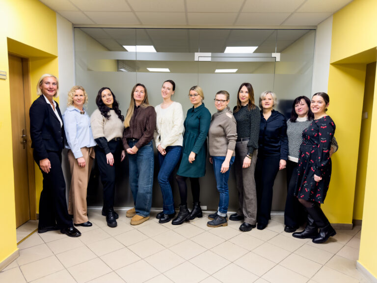 Nuotraukoje matyti vienuolika moterų, stovinčių greta viena kitos patalpoje su ryškiai geltonomis sienomis. Jos pozuoja bendrai nuotraukai ir žiūri į kamerą. Dauguma vilki dalykinio ar pusiau dalykinio stiliaus drabužius – švarkus, megztinius, sukneles, kelnes, avinčios žemakulnius ar aulinius batus. Už jų – stiklinė pertvara su matiniu viduriu, lubose matyti biuro tipo apšvietimas, grindys išklotos šviesiomis plytelėmis. Bendra nuotaika rami ir draugiška, moterys stovi artimai, kai kurios lengvai pasisukusios šonu.