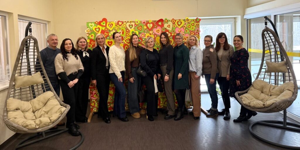 Nuotraukoje matyti grupė žmonių, stovinčių patalpoje prie spalvingos sienelės, dekoruotos daugybe spalvotų širdelių. Dauguma – moterys, taip pat vienas vyras. Visi pozuoja bendrai nuotraukai ir žiūri į kamerą. Jie vilki dalykinio ar pusiau dalykinio stiliaus drabužius. Patalpa šviesi, su dideliais langais. Iš abiejų pusių matyti pakabinamos pintos kėdės su minkštomis pagalvėmis. Atmosfera jauki ir draugiška, nuotrauka primena bendruomenės ar darbo kolektyvo susitikimą.