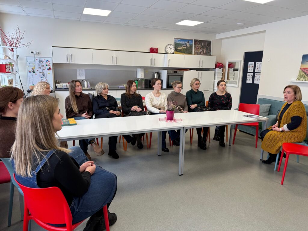 Nuotraukoje matyti susitikimas šviesioje patalpoje, primenančioje bendruomenės centrą ar mokymo klasę su virtuvės zona. Aplink stačiakampį stalą sėdi moterys, jos klausosi dešinėje pusėje sėdinčios ir kalbančios moters. Dalyvės vilki kasdienio ar pusiau dalykinio stiliaus drabužius, sėdi ant raudonų kėdžių. Ant stalo stovi nedidelis vazonėlis su augalu, padėta knyga ar užrašai. Fone matyti virtuvinės spintelės, laikrodis, paveikslai ant sienos. Nuotaika rimta ir susikaupusi, visos dalyvės dėmesingai stebi kalbančiąją.