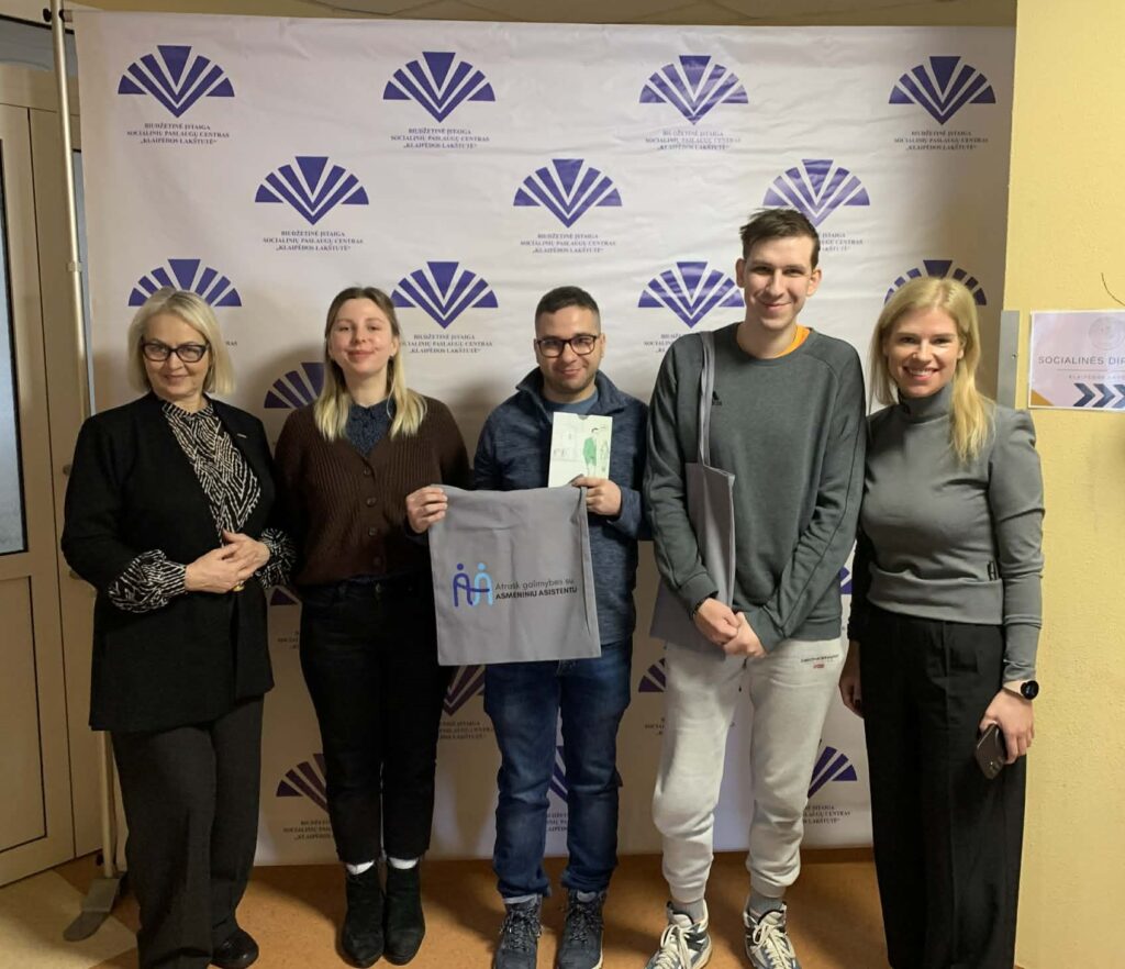 Prie foto sienelės stovi  5 žmonės ir pozuoja nuotraukai, šypsosi. Iš kairės: Socialinių paslaugų centro „Klaipėdos lakštutė" direktorė ir ANTA LSK regiono koordinatorė Danutė Daukantienė, ANTA lengvai suprantamos kalbos koordinatorė Laura Mikelevičiūtė, LSK vertintojas Karolis Juškėnas, LSK vertintojas Lukas Januškevičius, Socialinių paslaugų centro „Klaipėdos lakštutė" socialinių programų koordinatorė ir ANTA LSK regiono koordinatorė Justina Kubilienė. 