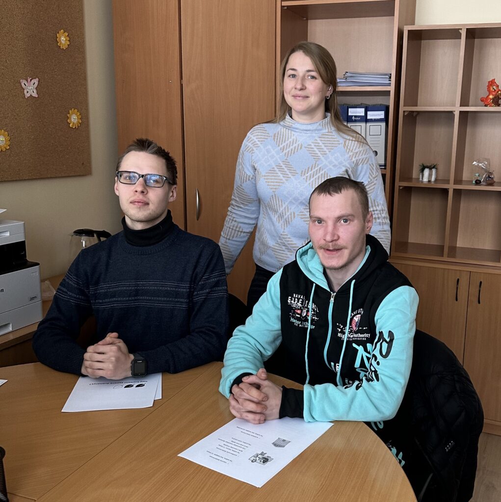 Kambaryje prie stalo sėdi du vyrai, o už jų sotvi moteris. Kambaryje prie sienos yra dvi komodos, spinta, ant sienos yra lenta. Nuotraukoje esanti moterys yra Panevėžyje dirbanti LSK regiono koordinatorė, atvejo vadybininkė Monika Bieliūnė. Prie stalo sėdintys vyrai yra LSK vertintojai Dovydas Baltramiejus Dirsė ir Dovydas Abromaitis. 