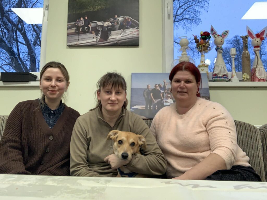 Ant sofos kambaryje sėdi LSK regiono koordinatorė, Sutrikusio intelekto žmonių globos bendrija „Šilutės Viltis“ pirmininkė Eglė Paulauskienė ir Lina Naujokaitė. Ant lovos taip pat sėdi mažas šuo. Ant sienos už sofos matyti paveikslai, taip pat yra langai, ant palangių sudėti dirbiniai.