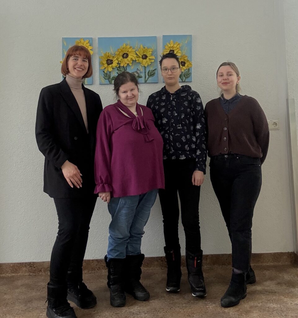 Prie sienos, ant kurios yra ryškių saulėgražų paveikslas, stovi 4 moterys: kairėje yra LSK regiono koordinatorė, Pagėgių SD rinkodaros specialistė Karolina Martišiūtė-El Oreibi, šalia jos dešinėje yra LSK vertintoja Jurgita Buivydaitė, LSK vertintoja Marija Amanovaitė, LSK koordinatorė Laura Mikelevičiūtė. Moterys pozuoja nuotraukai, šypsosi.