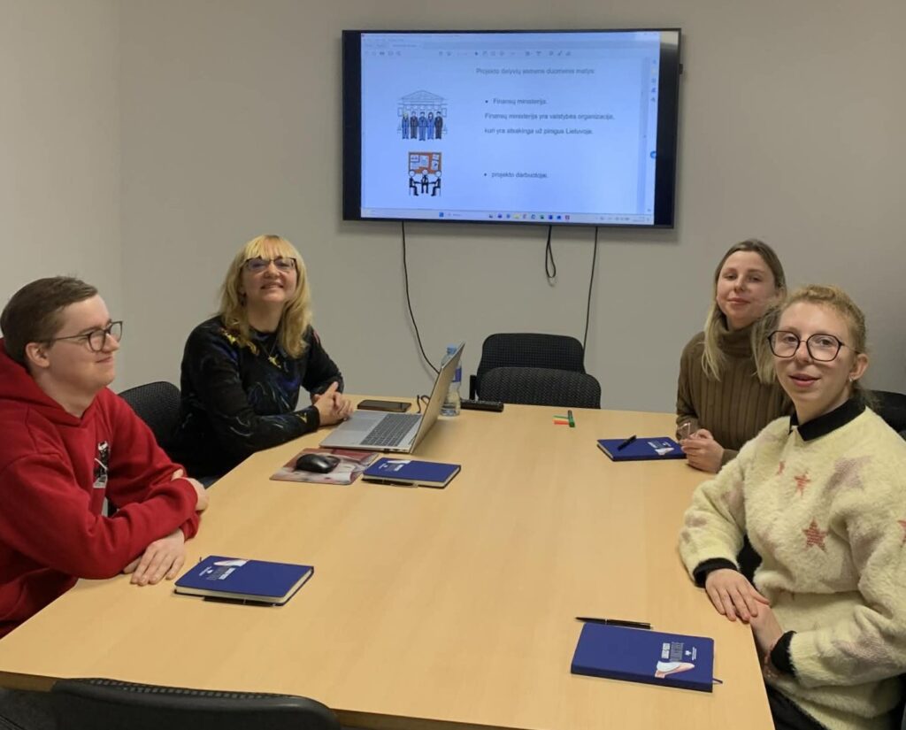 Salėje prie stalo sėdi keturi žmonės: LSK regiono koordinatorė Vilija Zeliankienė, LSK vertintojai Rytis Mitrius, Elzė Armonaitė ir LSK koordinatorė Laura Mikelevičiūtė. Ant stalo sudėtos knygos ir rašikliai, kompiuteris. Ant sienos yra pakabintas ekranas, kuriame yra rodomas lengvai suprantamos kalbos tekstas.