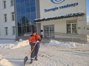 Žiemos metu prie administracinio pastato su užrašu „Tauragės vandenys“ stovi darbuotojas su ryškia oranžine atšvaitine darbo striuke, tamsiomis kelnėmis ir kepure. Jis laiko sniego kastuvą ir valo sniegą nuo tako priešais pastato įėjimą. Prie įėjimo matomi laiptai ir pandusas su turėklais. Aplink yra sniegas, šviečia saulė, o pastato stikliniai langai atspindi medžius.